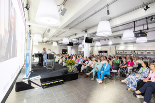 Eine Frau spricht auf einer Bühne vor einem Publikum in einem modernen Raum, umgeben von Pflanzen und großen Lampen.