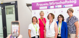 Das Bild zeigt eine Gruppe von Frauen, die lächelnd vor einem Banner des Frauen-Wirtschaftstags posieren. Eine Person macht ein Foto.