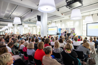 Das Bild zeigt eine große Gruppe von Menschen in einem modernen Veranstaltungsraum, die einer Präsentation aufmerksam folgen. Helle Lampen und eine freundliche Atmosphäre prägen den Raum.