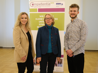 Das Bild zeigt drei Personen, die vor einem Banner stehen. Sie lächeln und scheinen an einer Veranstaltung oder einem Treffen teilzunehmen.