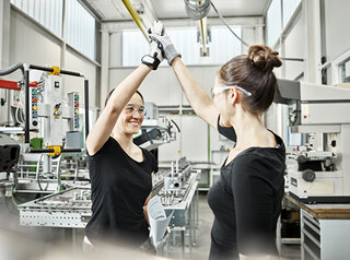 Zwei Frauen in Schutzkleidung geben sich im modernen Fertigungsraum ein High-Five. Die Umgebung ist industriell, mit Maschinen und Werkzeugen.