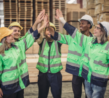 Auf dem Bild sind sechs Arbeiter in Sicherheitskleidung und Helmen zu sehen, die sich zu einem High-Five zusammenschließen. Alle lachen und wirken motiviert.